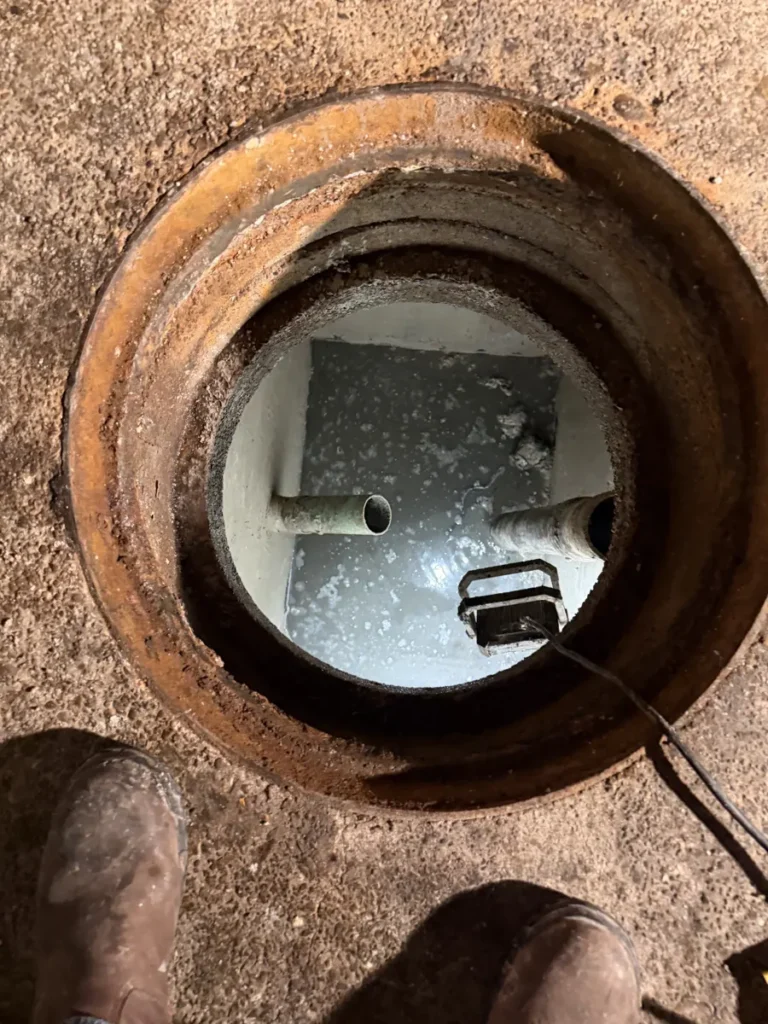 Open manhole of an in-ground grease trap, with two pipes and a work light in its coated interior below