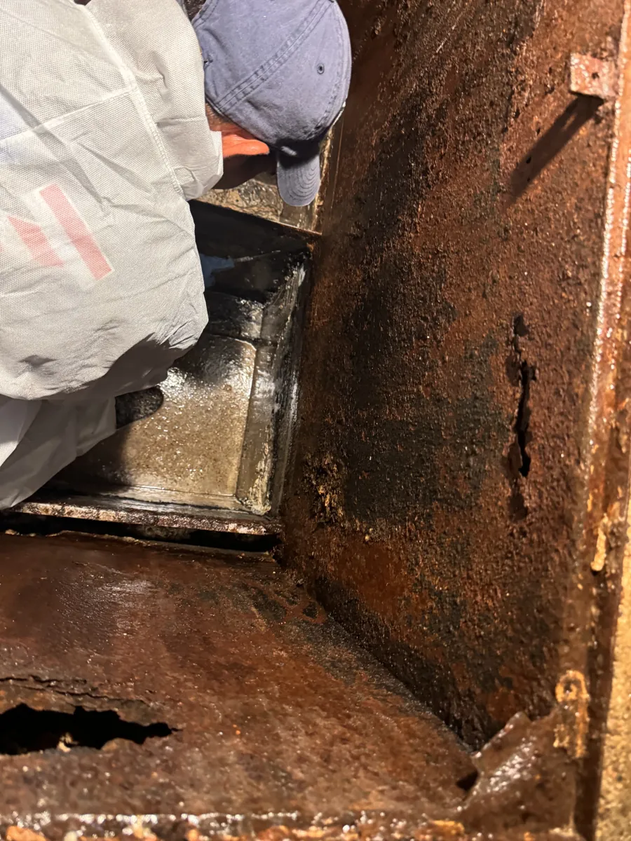 Worker checking a steel grease trap whose wall is rusted through, with holes along the bottom edge